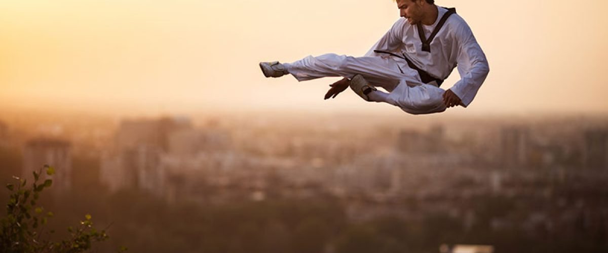 Teakwondo fighter performing high kick while being in mid air at sunset in fly-kick pose.
